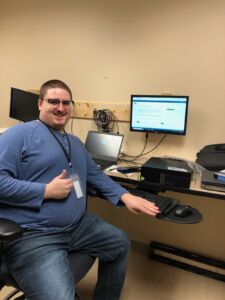 Photo shows a men in a blue long sleeve shirt. He is sitting in front of a computer and giving a thumbs up to the camera. He is wearing a company ID on a lanyard around his next