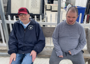 Image shows two men seated on a bench. One is wearing a baseball hat.