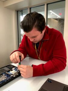 Photo shows a man in a red fleece jacket working on repairing a laptop