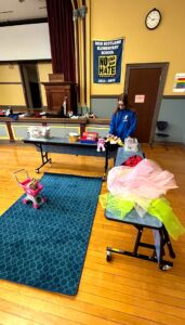 Photo shows a woman wearing a blue YMCA staff jacket. She is standing behind a table, with various toys around the room. A banner on the wall behind her reads, "New Scotland Elementary School. No Place for Hate. 2022-2023."