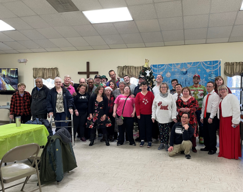 Over two dozen people pose for a photo. A winter wonderful backdrop, Christmas tree, and cross can be seen behind the group.