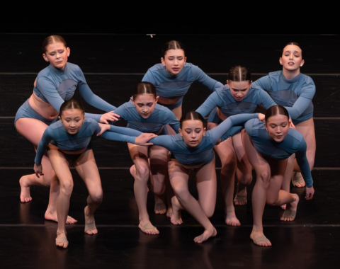 Eight dancers are stepping forward during a performance. They are standing over each other, their facial expressions are full of emotion. They are wearing blue costumes.