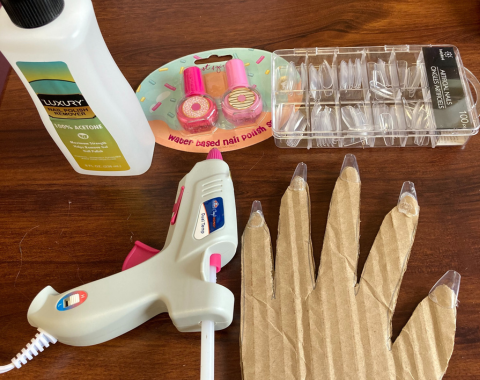 A cardboard cut-out of a hand has clear artificial nails glued on it. It’s on a table top next to a hot glue gun, a package of artificial nails, a package of water-based nail polish, and a bottle of acetone.
