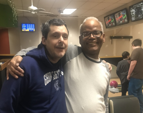 Two men stand with their arms around each others shoulders. They are both smiling. A bowling alley can be seen behind them.