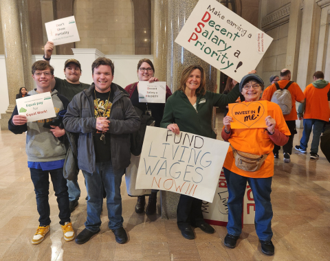 A group of six people pose for a photo. They are holding signs that say things like "Make Earning a Decent Salary a Priority" and "Fund Living Wages Now!" One of the people is Living Resources CEO Elizabeth Martin.