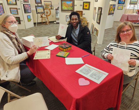 Three people are seen coloring at a table covered in a red table cloth. They are holding up their creations and smiling for the camera.