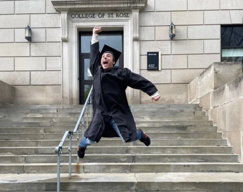 A student wearing a cap and gown is jumping in the air in front of an academic building. Words above the building's door read "College of St. Rose."