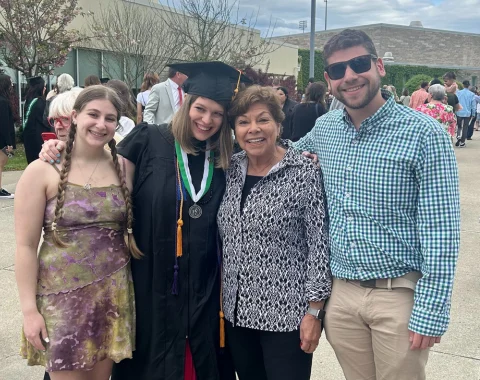 Four people pose for a photo. One of the women in the middle is wearing a black graduation cap and gown.