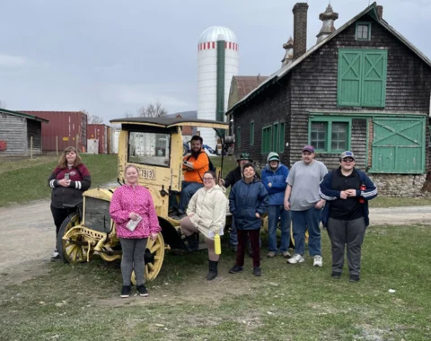 Nine people pose for a photo in front of a brown barn with green trim. A silo is seen in the background. One of the people is sitting in an old yellow farm vehicle.