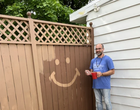 A man poses for a photo next a a fence he is painting. He's used the paint to put a smiley face on the fence.