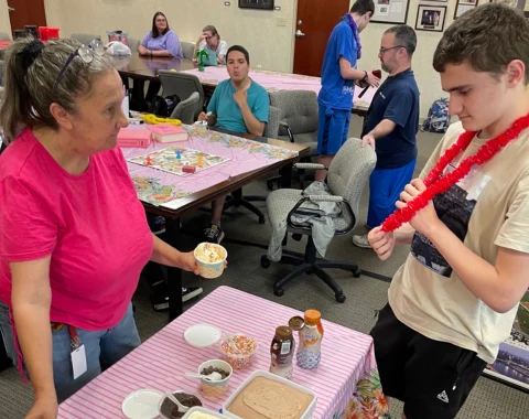 A woman and a boy are at the make your own sundae bar. Other program participants are seen in the background enjoying the party.