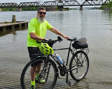 A man poses for a photo next to his bicycle near a body of water. A bridge is seen in the background.