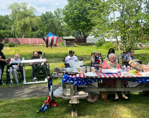 Seven people sit outside for a cookout. An American flag-decorated tablecloth and balloon are with them.