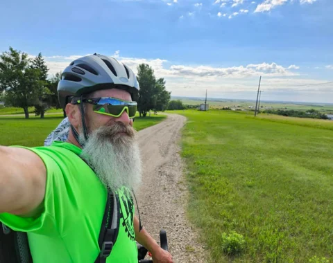 A man poses for a selfie on a long dirt road. Green fields can be seen on either side of the road.