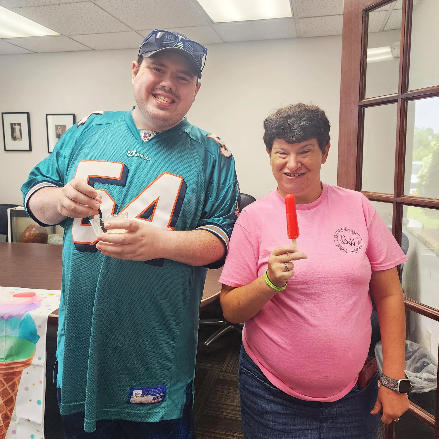 Ice Cream Social 4 A man in a miami dolphin's shirt stands next to a woman in a pink shirt smiling. The man is holding a cup of ice cream. Th woman holds a red popsicle.