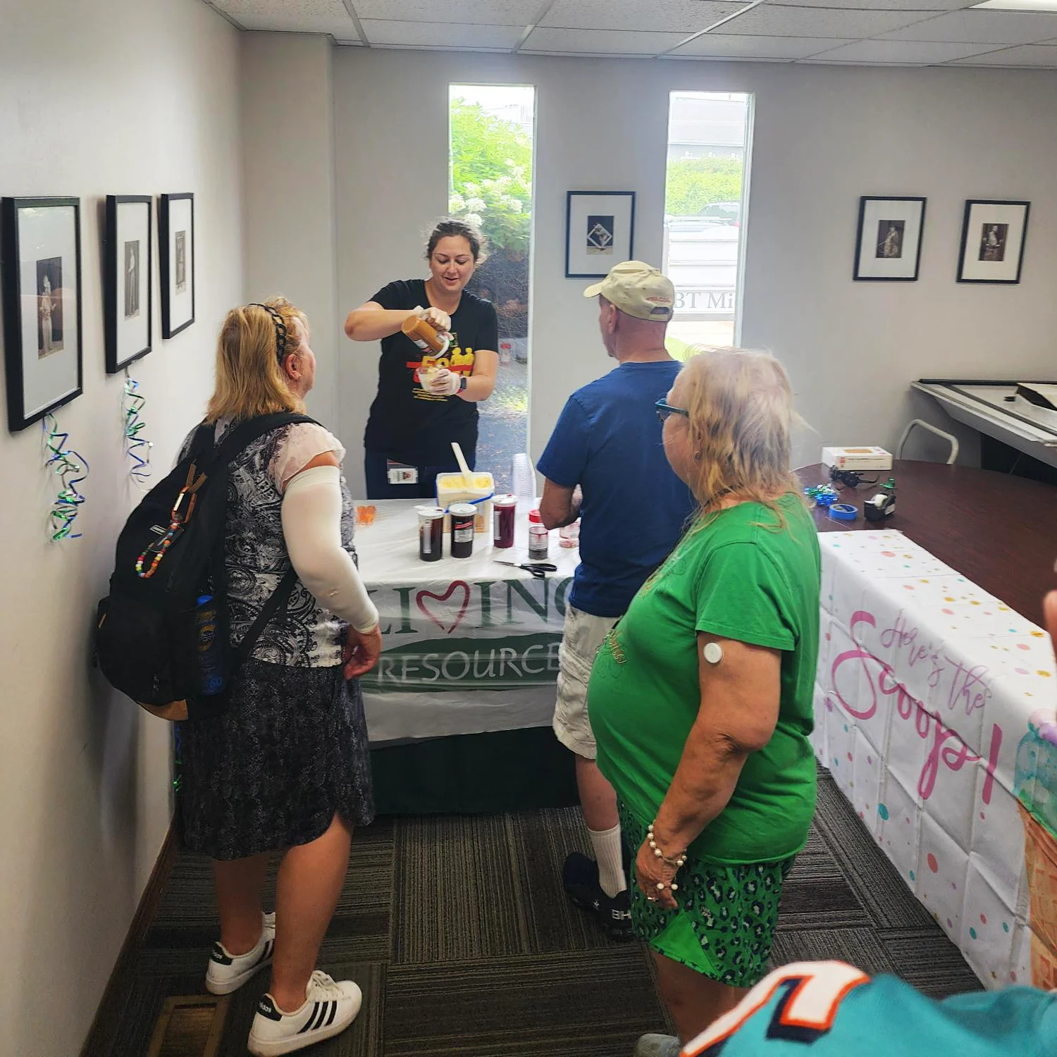 Ice Cream Social 5 A woman stands behind a table with ice cream and toppings on it, pouring caramel onto a sundae. 3 people stand in line