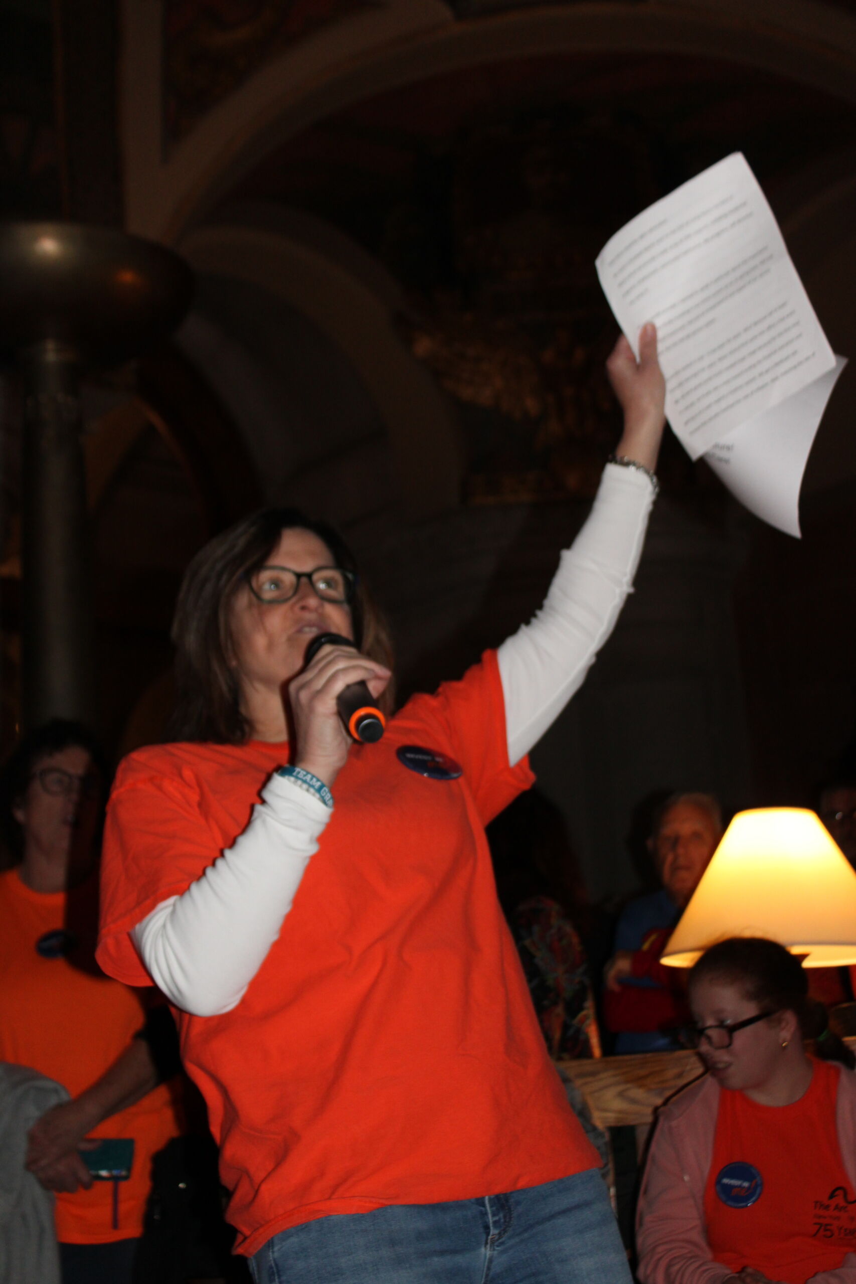 A woman in an orange shirt holds a piece of paper in one hand held high and holds a microphone in the other hand to her mouth. She is giving a speech.
