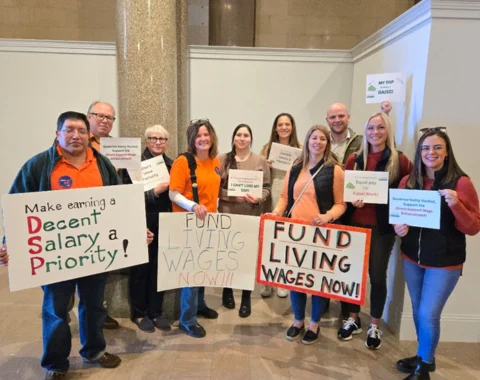 a group of people stand holding signs advocating for better wages for direct support professionals. Example signs read "Fund Living Wages Now" and "Make Decent Salary a Priority".