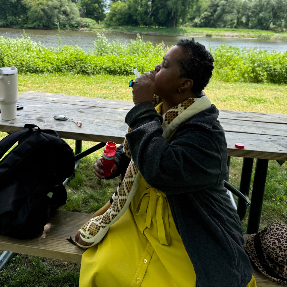 A woman in a yellow dress and black jacket with a stuffed yellow snake around her neck sits facing the left and blows bubbles on a park bench.