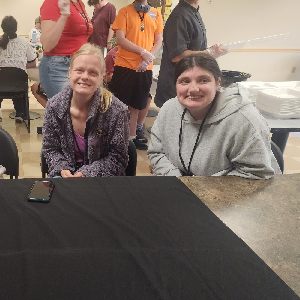 2 Two female students pose for a photo at a table with a black tablecloth on it.