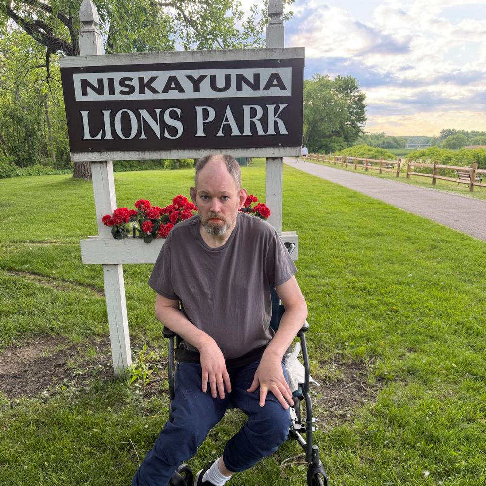 Image shows a man sitting in a wheelchair in front of a sign that says Nisayuna and then Lyons Park in large white letters. He is wearing a grey tshirt, jeans, and black shoes.