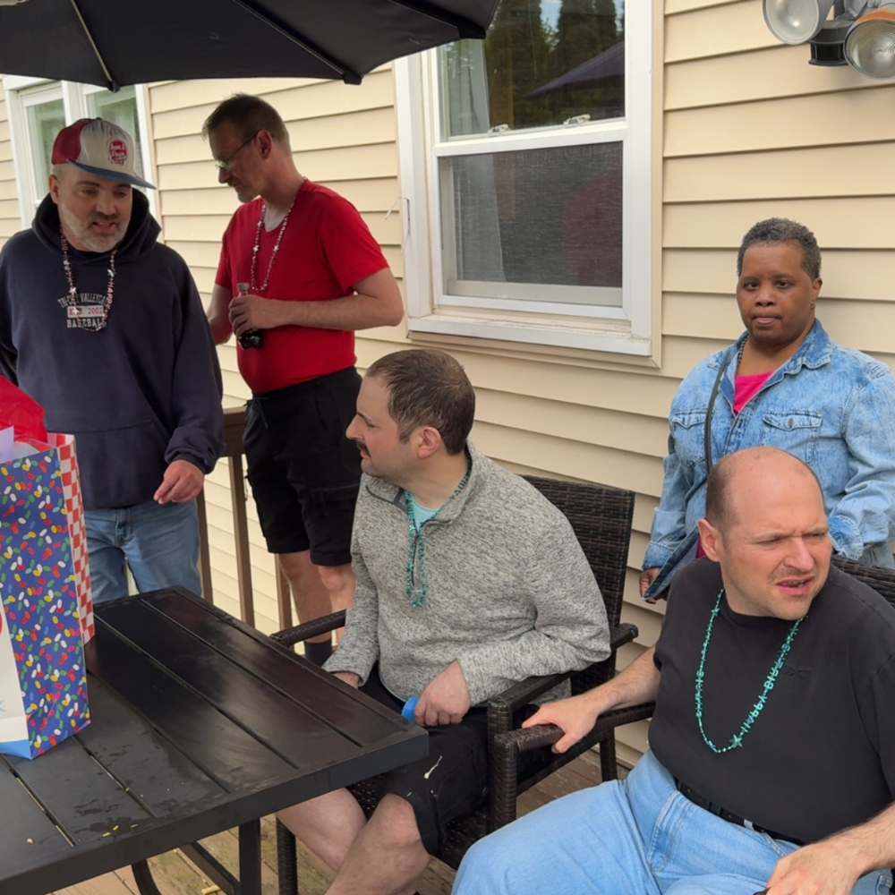 Image shows individuals in living resources residential program sitting and standing around a patio table with a black umbrella.