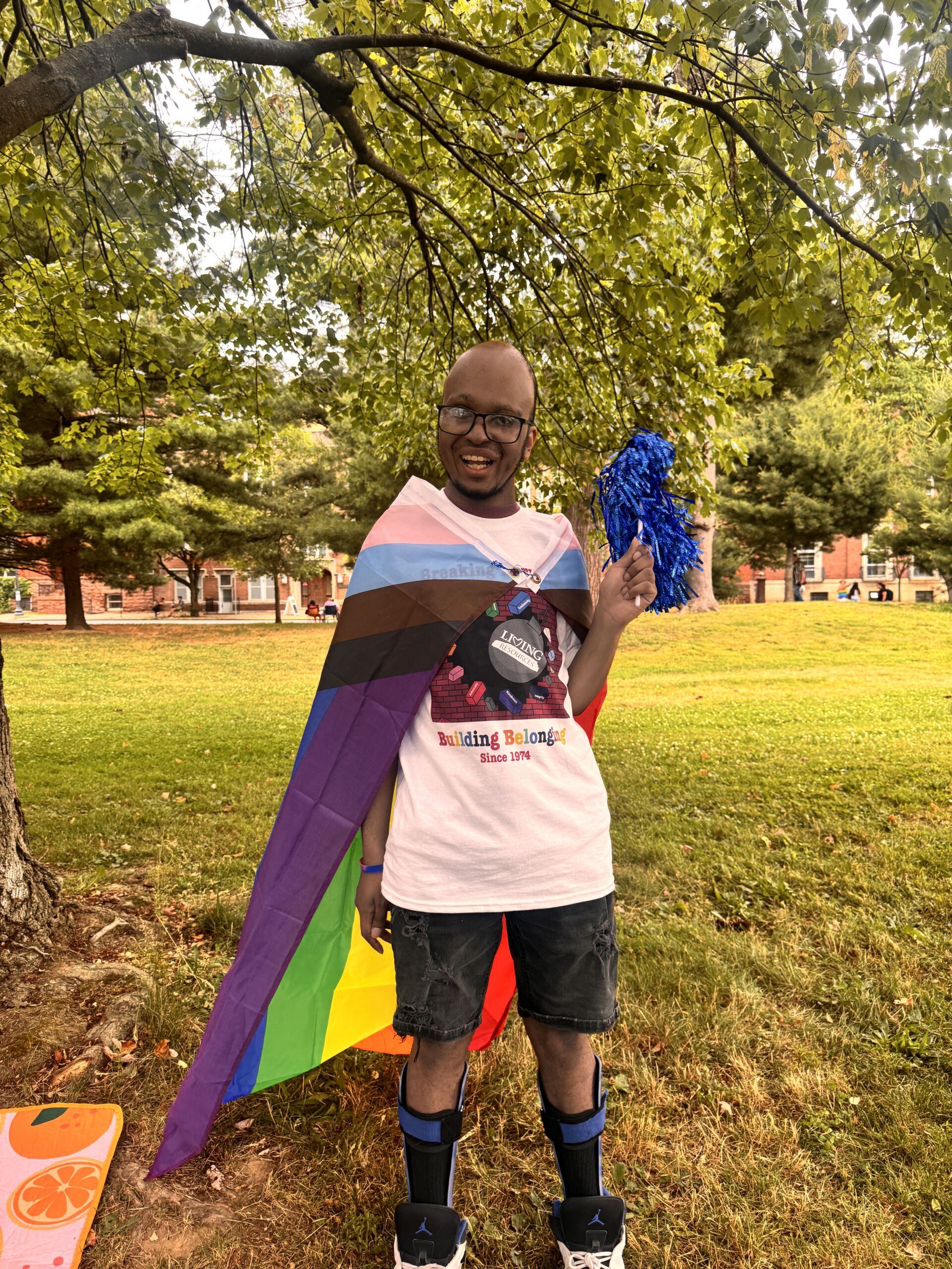 IMG_2051 1 Image is of an individual supported by living resources at the Albany pride parade. He is wearing a shirt that says breaking barriers building belonging. He has a pride flag wrapped around his body like a cape and is smiling at the camera.