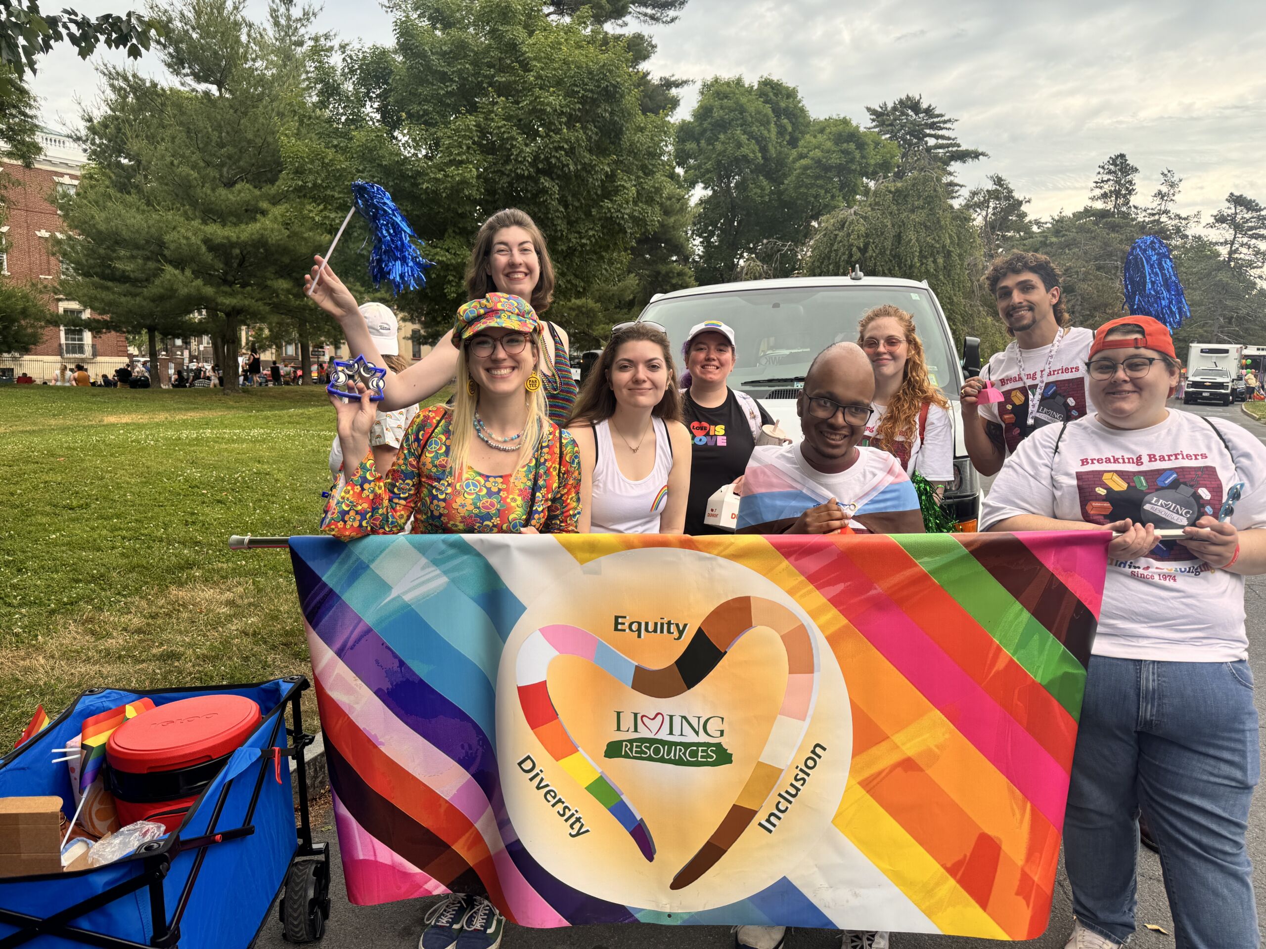 IMG_2055 1 Image is of Living Resources staff and supported individuals getting ready to march in the albany capital pride parade. They stand behind a horizontal banner that features both the LGBTQ+ pride flag colors along with the disability pride flag colors.