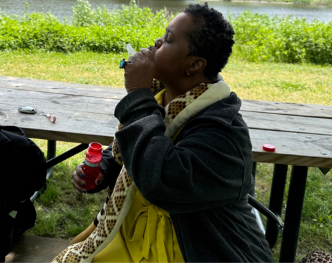 A woman in a yellow dress and black jacket with a stuffed yellow snake around her neck sits facing the left and blows bubbles on a park bench.