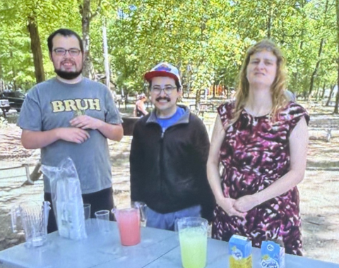 Image shows three individuals supported by Living Resources' DCOP holding a lemonade stand in a park. They are standing at a grey folding table with pitchers of pink lemonade and regular lemonade. There are cups on the table and the three individuals smile at the camera.