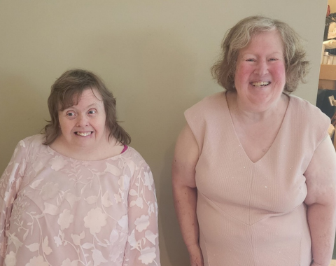 Two women smiling warmly while posing side by side in front of a neutral-colored wall. The woman on the left wears a light pink long-sleeved floral lace top, and the woman on the right wears a sleeveless light pink v-neck dress.
