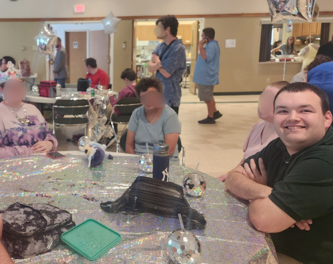 A young man sitting at a table covered in a shiny silver tablecloth, smiling directly at the camera with his arms crossed. Nearby at the table and in the background, other event attendees sit and stand around a brightly lit community hall decorated with silver star balloons and disco ball drink cups.