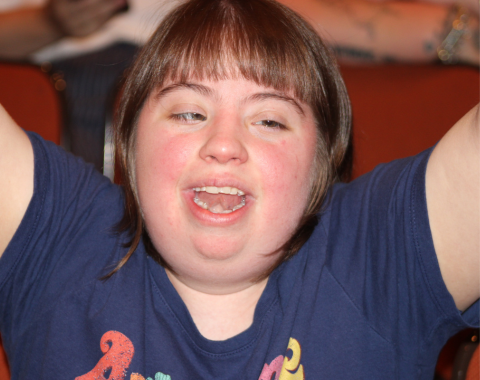 A woman with Down Syndrome smiles for the camera with her hands raised in excitement.