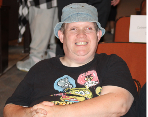 Day Services Awards 6 A man in a black shirt and gray bucket hat smiles for the camera while sitting in his seat in an auditorium.