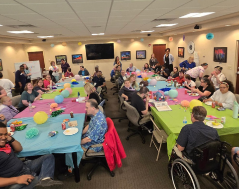 A large room is decorated for a party. Many party-goers fill the room and sit at colorfully decorated tables.