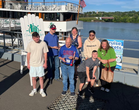 A group of seven people posing together outdoors on a sunny dock next to a river. In the background is the Dutch Apple Cruise boat and a water view. An oversized white wooden Adirondack chair bearing the Dutch Apple logo stands to the left of the group.