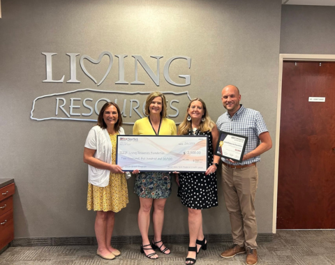 Four people stand in front of a gray wall with this living resources logo on it in silver. The four people are holding a large check as First New York Federal Credit Union presents Living Resources with a $2,500.00 donation.