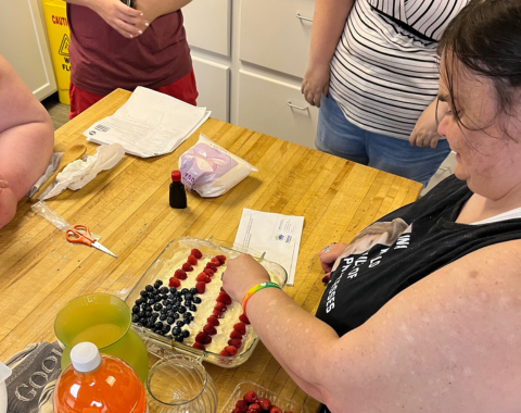An overhead shot of an individual carefully placing fresh raspberries in neat lines onto a frosted sheet cake to create the red stripes of an American flag, while a cluster of blueberries forms the blue star field in the corner.