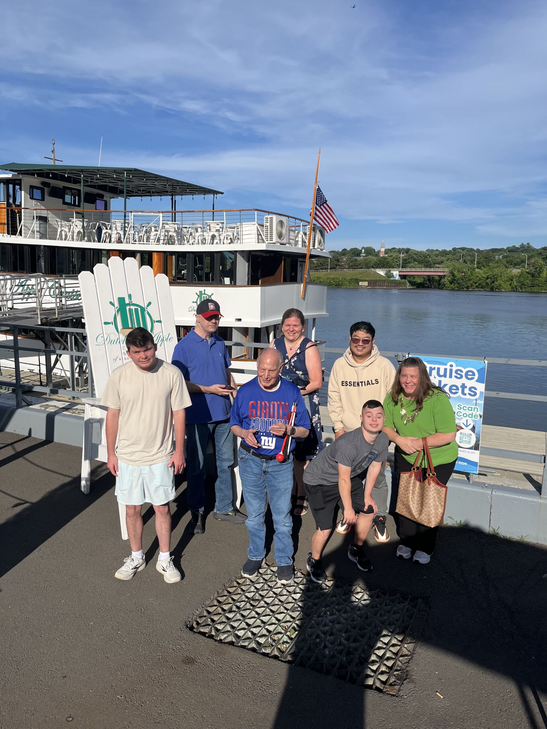Group Photo A group of seven people posing together outdoors on a sunny dock next to a river. In the background is the Dutch Apple Cruise boat and a water view. An oversized white wooden Adirondack chair bearing the Dutch Apple logo stands to the left of the group.