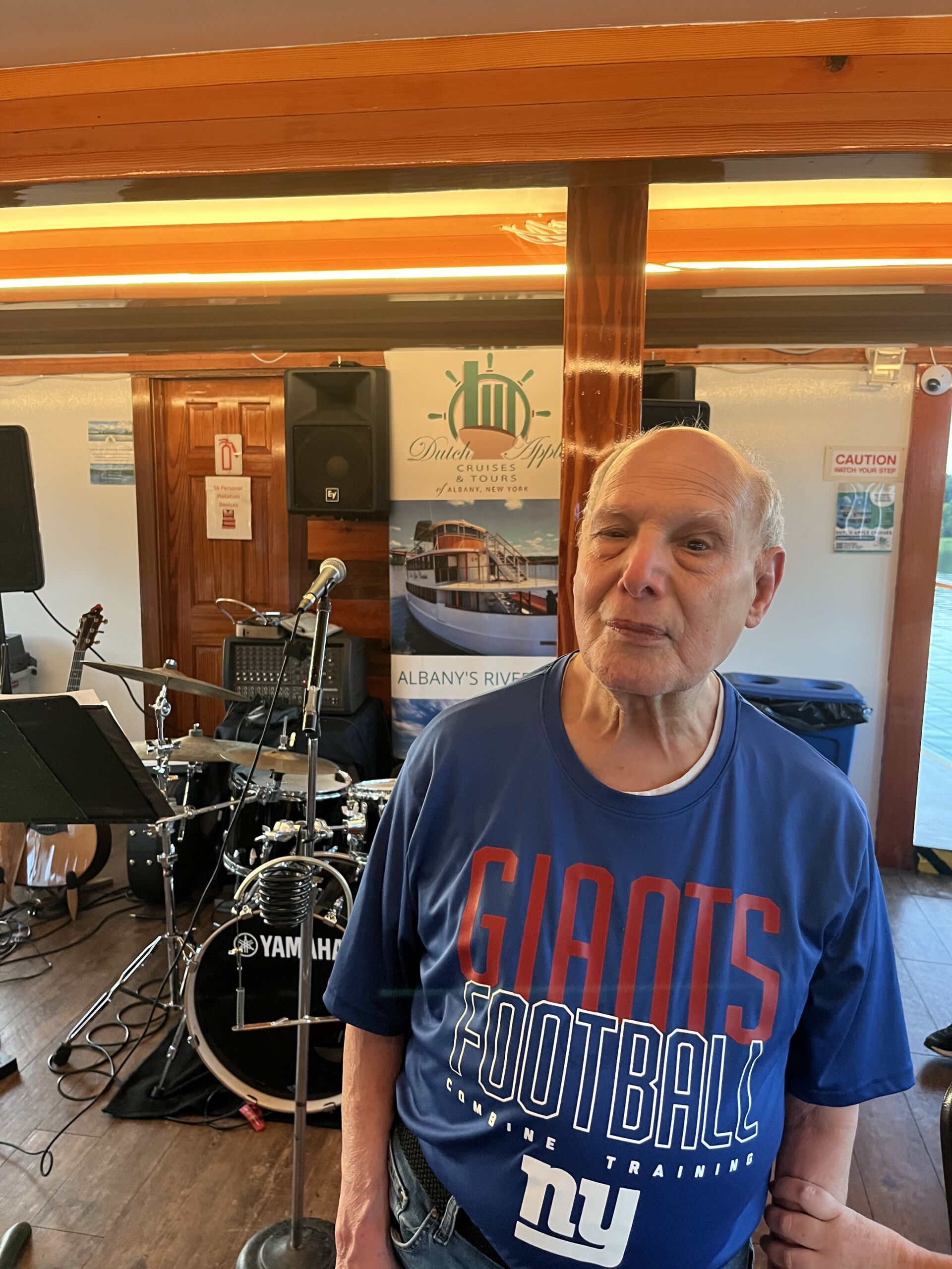 Al Dutch Apple Cruise A close-up indoor portrait of an older man smiling while wearing a blue New York Giants Football t-shirt. Behind him, a drum kit, audio equipment, and a Dutch Apple Cruises & Tours banner are visible inside the boat's interior.