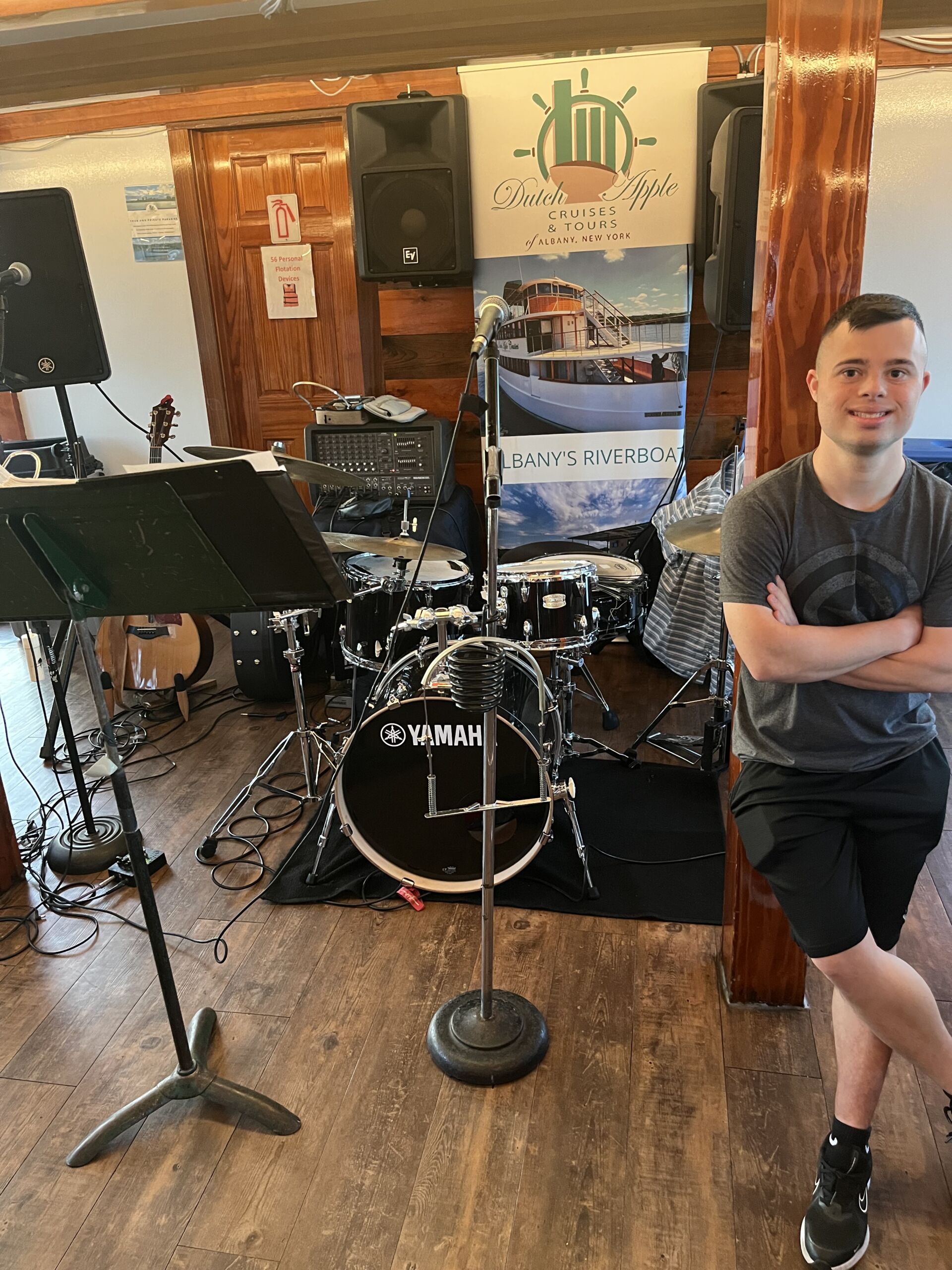 Joe Dutch Apple Cruise A young man wearing a grey t-shirt and black shorts leaning against a wooden pillar with his arms crossed, smiling at the camera. In the background is an indoor stage setup featuring a Yamaha drum set, a microphone stand, and a musical equipment array inside a cruise boat.