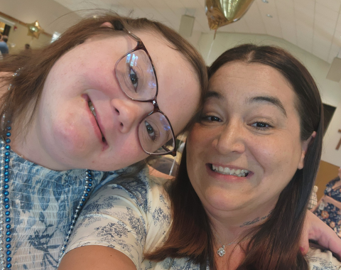 A close-up selfie of two women smiling with their heads tilted together. The woman on the left wears dark-framed glasses, a blue patterned top, and blue beaded necklaces. The woman on the right has shoulder-length brown hair, a silver necklace, and a light-colored patterned shirt.