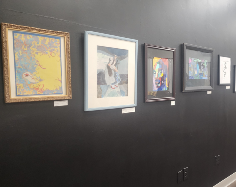 88 Unbound Show A perspective shot looking down a long, dark wall in an art gallery setting with dark wood floors. A linear gallery display features over ten distinct framed artworks of various sizes, styles, and frame colors (gold, light blue, dark wood, black). The artwork ranges from colorful abstract paintings and portraits to black-and-white line drawings and detailed illustrations, each with a small white label mounted below it.