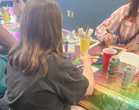 888 unbound party A person with shoulder-length brown hair sitting on a black folding chair, viewed from behind. They are wearing a dark grey t-shirt and beige shorts, leaning forward to paint a small object at a table covered with a iridescent rainbow plastic tablecloth. Across the table, another person wearing a striped shirt and a red name tag works on their own painting project. The table is filled with art supplies, including yellow cups holding paintbrushes, paint palettes with colorful dollops, red cups, and soda cans.