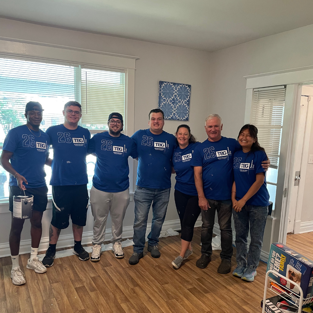 Bonadio Group Shot Seven volunteers stand side-by-side with arms wrapped around one another in an indoor room with light wood floors. All wear matching royal blue t-shirts with "26 Purpose Day" and "The Bonadio Group" printed on the front. One man on the left holds a paint container, and paint smudges are visible on several of their shirts and pants.