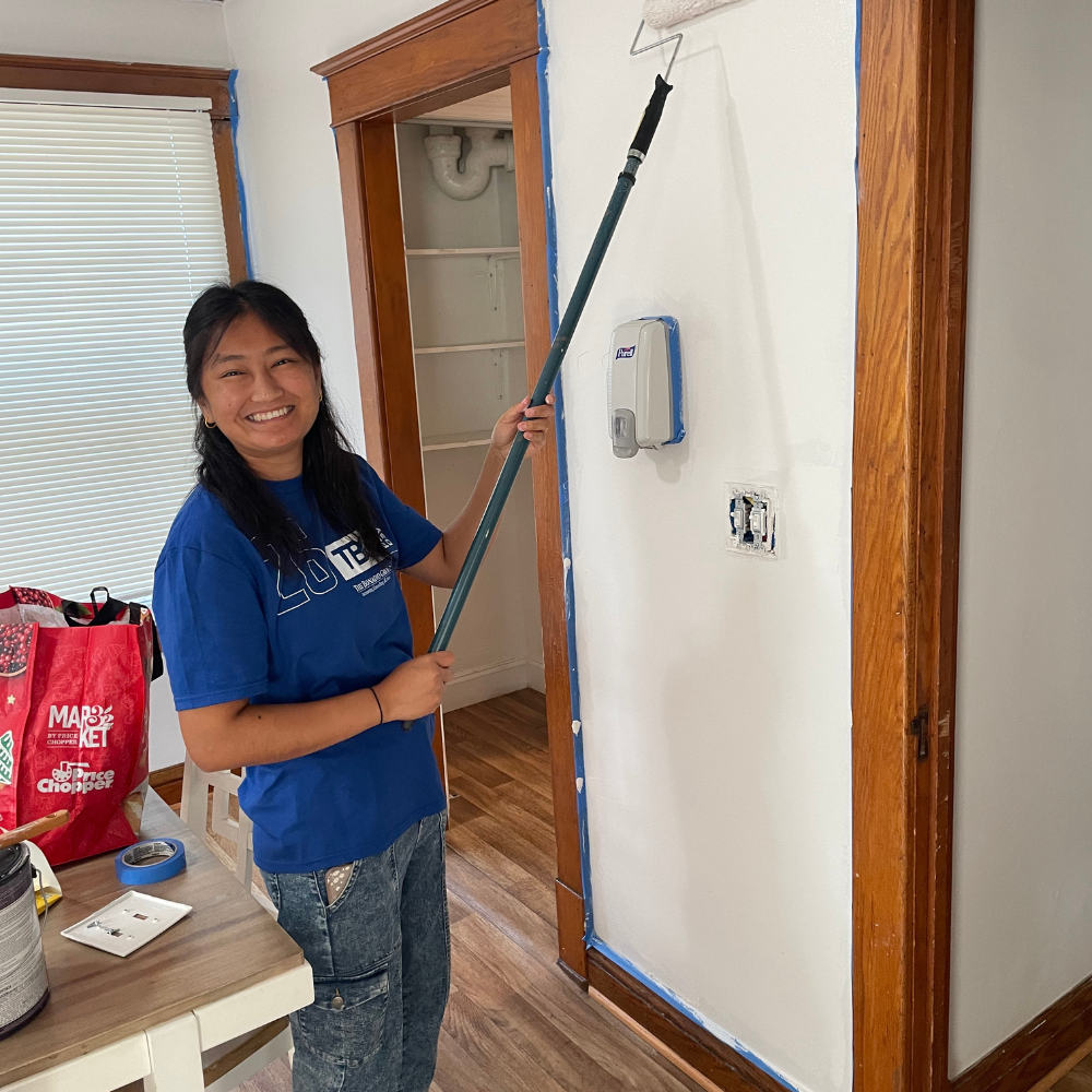Bonadio Volunteer 1 A smiling woman with dark hair stands inside near a doorway, wearing a royal blue Bonadio t-shirt and grey jeans. She holds a long extension pole with a paint roller, applying fresh white paint to a section of wall framed in wood trim taped off with blue painter's tape.