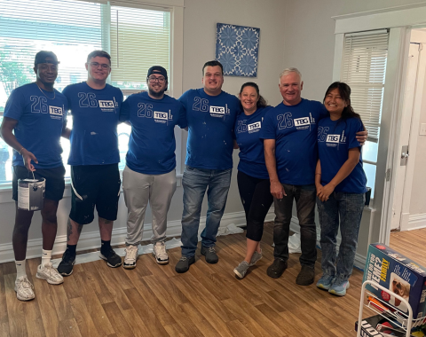 Group shot of seven volunteers smiling in a row inside an indoor room, all wearing matching bright blue t-shirts featuring the logo for The Bonadio Group. They are taking a break from painting, with one person holding a paint can on the far left.