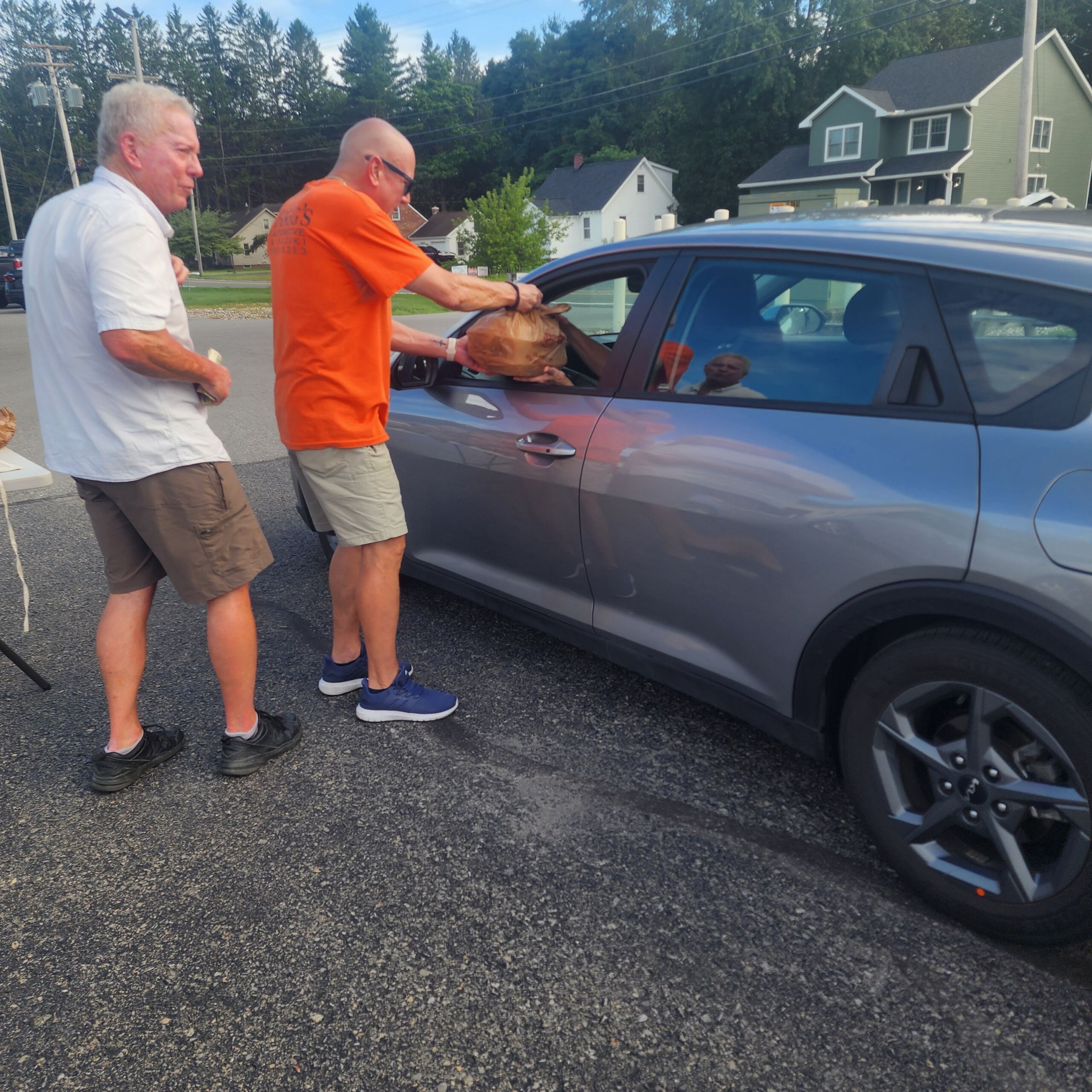 Foggs BBQ 1 Two volunteers standing next to a grey crossover SUV in a drive-thru line, with one man in an orange shirt handing a brown paper bag of Brooks BBQ food to the driver through the window.