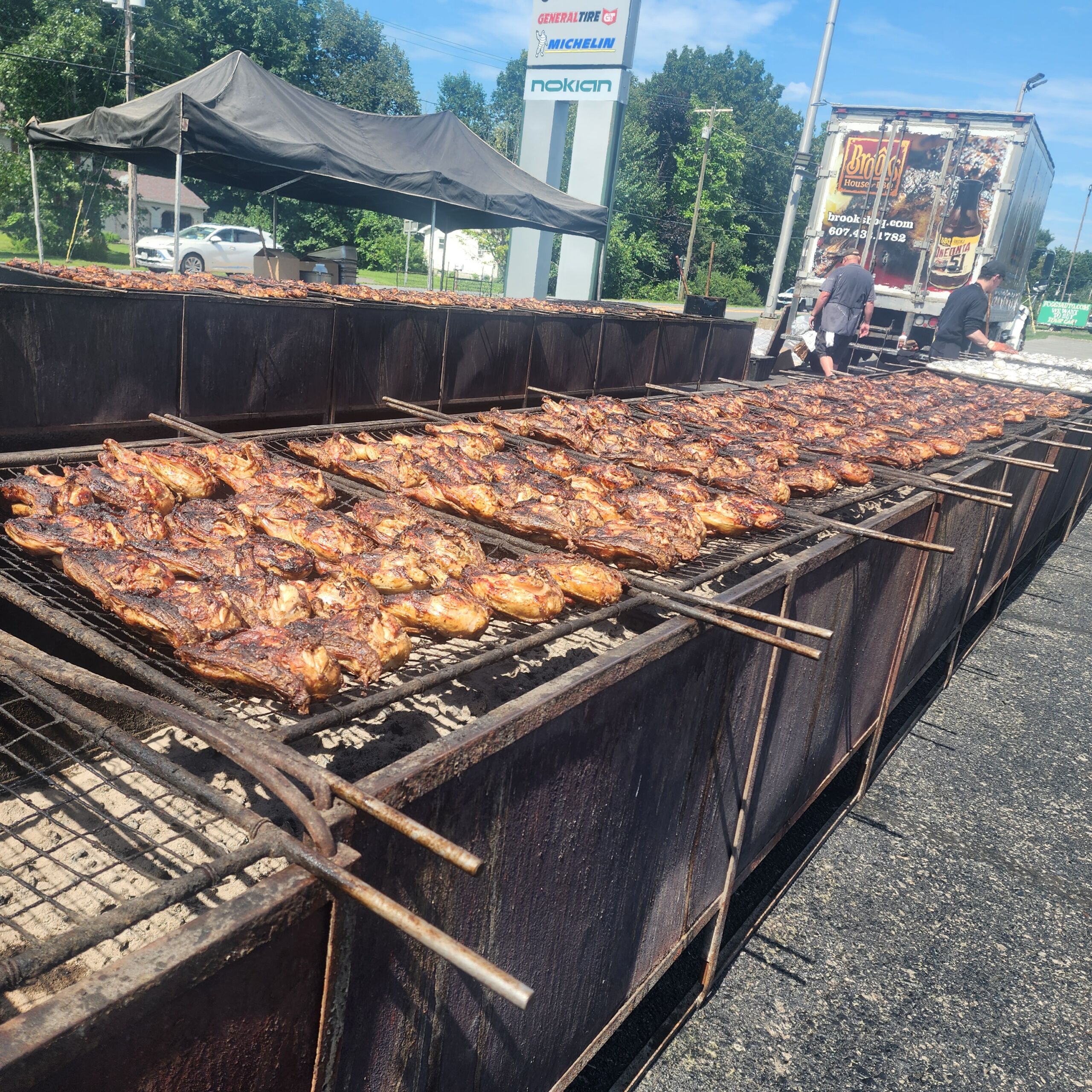 Foggs BBQ Chicken Rows of Brooks BBQ chicken quarters sizzle over large charcoal pit grills outdoors, with a dark canopy tent, a Brooks House BBQ catering truck, and tire dealership signage visible in the background.