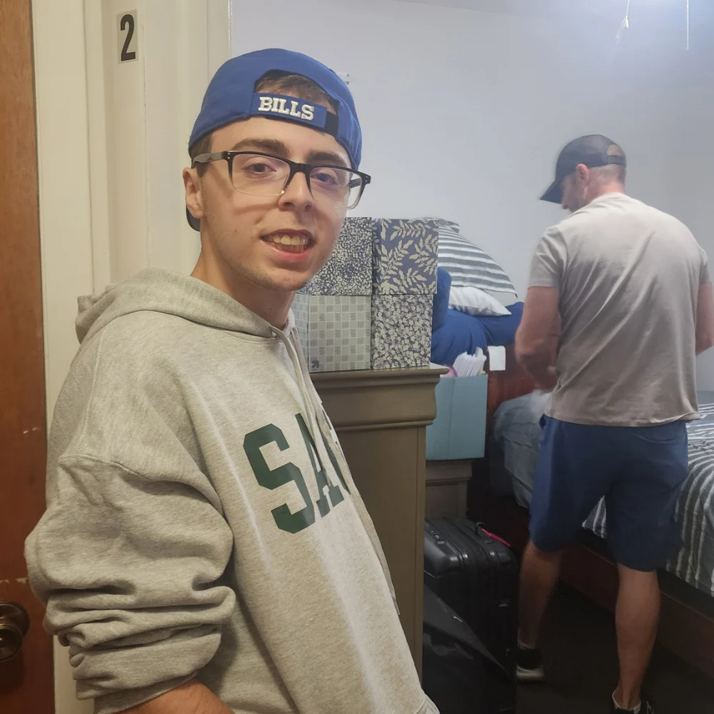 Move in Day 1 A young man wearing glasses, a blue "BILLS" baseball cap turned backward, and a grey hoodie smiles at the camera inside a bedroom. In the background, another person works near a bed surrounded by stacked decorative storage boxes, suitcases, and pillows.